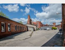 Industriehof mit Backsteingebäuden, großem gepflastertem Hof und mehreren parkenden Fahrzeugen. Im Hintergrund sind ein Schornstein und weitere Backsteingebäude mit roten Dächern zu sehen, umgeben von bewölktem Himmel.