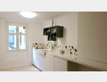 Eine minimalistisch gestaltete Küche mit weißer Einrichtung, Waschbecken und Arbeitsflächen. Die Wand ist mit einer Terrazzo-Optik gestaltet, ein kleines Fenster lässt Tageslicht herein, und ein hängendes Regal bietet Stauraum.