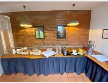 Ein Buffet-Aufbau in einem Raum mit einer Ziegelsteinwand, dekoriert mit zwei hängenden Lampen. Auf dem Tisch stehen Gläser, Karaffen mit Getränken, Wasserflaschen sowie Körbe mit Snacks, dazu eine silberne Teemaschine auf der rechten Seite.