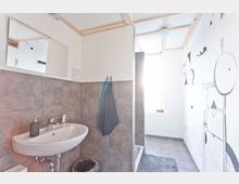 Kleines, schlichtes Badezimmer mit grauen Fliesen, einem Wandwaschbecken, einem kleinen Spiegel und einer teils offenen Duschkabine. Die weiße Wand rechts ist mit abstrakten, schwarzen Grafiken verziert, und ein Fenster im Hintergrund lässt Tageslicht herein.