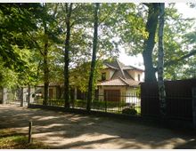 Zweistöckiges Haus mit gelbem Putz und einem Dach aus roten Ziegeln, umgeben von Bäumen und einem Metallzaun, gelegen an einem unbefestigten Weg in einer waldreichen Umgebung.