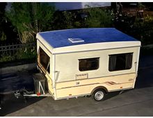 Ein heller, kompakter Wohnwagen steht am Straßenrand, umgeben von Pflanzen und einem Metallzaun im Hintergrund. Der Wohnwagen hat ein flaches Dach, rechteckige Fenster mit Vorhängen und eine Anhängerkupplung an der Vorderseite.