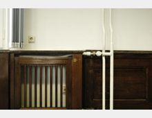 Holzvertäfelte Wand mit einem Heizkörpergitter und sichtbaren weißen Rohren, daneben eine Steckdose. Die Wand ist weiß gestrichen, und ein Fenster mit geschlossenen Rollos befindet sich links im Bild.
