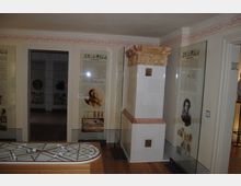 Ein Museumsraum mit historischen Texttafeln und Bildern hinter Glasvitrinen, einem verzierten weißen Kachelofen in der Ecke und einem Holzmodell mit geometrischen Linien auf einem Tisch im Vordergrund. Der Raum hat eine helle Wandgestaltung mit roten Ornamenten entlang der oberen Kante.