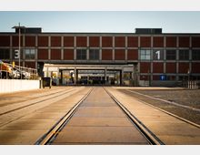Industriekomplex mit einer breiten Zufahrtsstraße, eingebetteten Schienen und einem Backsteingebäude im Hintergrund. Auf der linken Seite sind geparkte Autos und Abfallcontainer zu sehen, während Schilder und Durchgänge die Bereiche 1 und 3 markieren.