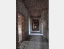 Korridor in einem verlassenen Gebäude mit unverputzten Ziegelwänden, einfachen Holzböden und einer Decke aus sichtbaren Schilfrohrmatten. Türöffnungen führen in weitere leere Räume.