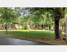 Eine eingeschossige, historische Backsteinfassade mit großen Fenstern, umgeben von einer gepflegten Grünfläche mit Rasen und zahlreichen Bäumen. Im Vordergrund verläuft eine gepflasterte Straße mit einem niedrigen Metallgeländer.