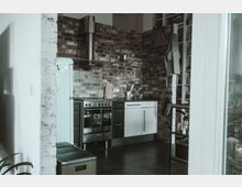 Moderne Küche mit Backsteinwänden, einem silbernen Herd mit Dunstabzugshaube, einer weißen Kühl-Gefrierkombination und weißen Unterschränken mit schwarzer Arbeitsplatte. Im Hintergrund befinden sich Regale sowie ein kleiner Esstisch.