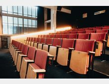 Reihen von Sitzplätzen mit roten Polstern und Holzlehnen in einem kleinen Auditorium. Große Fenster lassen natürliches Licht herein, während eine beleuchtete Wand im Hintergrund den Raum erhellt.
