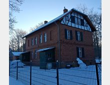 Backsteinbau eines zweigeschossigen Hauses mit Fachwerkelementen im oberen Giebelbereich, umgeben von einem verschneiten Gelände und einem Metallzaun. Im Hintergrund sind kahle Bäume und ein klarer Himmel zu sehen.