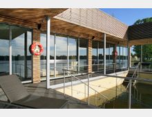 Modernes Gebäude mit Holzfassade und großen Glasfronten, das an einem Gewässer liegt. Im Vordergrund sind ein Geländer, Treppen und ein Liegestuhl zu sehen; Rettungsringe hängen an den Wänden, und die Umgebung zeigt Bäume und leicht bewölkten Himmel.