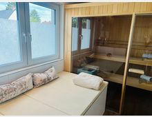 Ein moderner Wellnessbereich mit einer Holzsauna hinter einer Glastür, einem beigen gepolsterten Ruheliegeplatz mit Kissen und Handtuch sowie zwei Fenstern mit Sichtschutzfolie, die Tageslicht hereinlassen.