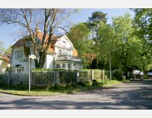 Ein zweigeschossiges Wohnhaus mit weißer Fassade und rotem Satteldach, umgeben von Bäumen und einem Metallzaun. Das Haus steht an einer Straßenecke in einer grünen, ruhigen Umgebung; im Vordergrund sind ein Straßenlaternenmast und ein Anhänger mit einem Boot zu sehen.