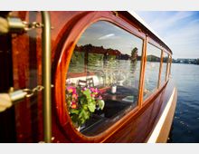 Holzboot mit verglasten Fenstern, die den Blick auf den elegant eingerichteten Innenraum mit Blumen und einem gedeckten Tisch freigeben; im Hintergrund ein Fluss mit Bäumen am Ufer und einer Brücke in der Ferne.
