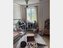 Ein helles Kinderzimmer mit Holzfußboden, einem niedrigen Kinderbett auf der linken Seite und einem Kletter- und Spielbereich mit Schaumstoffelementen in der Mitte. Im Hintergrund steht ein Regal mit Kleidungsstücken, Spielzeug und Dekoration neben einem Fenster mit hellen Vorhängen, durch das Tageslicht einfällt.