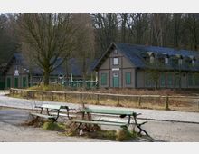 Holzgebäude mit grünen Fensterläden und einem Schieferdach in ländlicher Umgebung, umgeben von kahlen Bäumen. Im Vordergrund befinden sich zwei grüne Bänke auf einem sandigen Weg nahe einer gepflasterten Fläche.