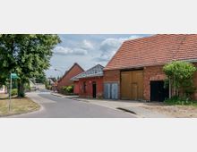 Straßenszene in einem ländlichen Dorf mit einer asphaltierten, leicht kurvigen Straße, die von Backstein- und Putzgebäuden mit roten Ziegeldächern gesäumt ist. Links steht ein großer Baum und ein Straßenschild, während im Hintergrund weitere Wohnhäuser und Grünflächen zu sehen sind.