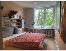 Modernes Schlafzimmer mit Doppelbett und rosafarbener Tagesdecke, wandmontierten Regalen und großformatigen Bildern an der Wand. Ein großes Fenster mit Heizkörper darunter bietet Blick auf grüne Bäume und lässt viel Tageslicht in den Raum.