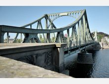Grüne Stahlbrücke mit bogenförmigen Verstrebungen über einem Fluss, mit steinernen Verzierungen am Brückengeländer und blauem Himmel im Hintergrund.