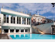 Außenansicht eines Hotels mit der Aufschrift „Inselhotel“ auf dem Dach. Im Vordergrund befindet sich ein rechteckiger Pool, umgeben von einer Holzterrasse, während eine Treppe rechts zu einer oberen Ebene mit Geländern führt.