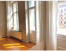 Heller Wohnraum mit großen, bodentiefen Fenstern, weißen Vorhängen und Holzdielenboden. Außenfassaden von Altbaugebäuden sind durch die Fenster sichtbar.