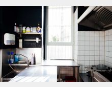 Eine kleine Edelstahlküche mit Waschbecken, Wandregalen, weißen Fliesen und einem großen Fenster mit Gitter. Links befinden sich Reinigungsmittel und rechteckige Arbeitsflächen, rechts eine große Dunstabzugshaube und ein Herd.