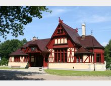 Historisches Gebäude im Fachwerkstil mit roten Holzverzierungen und cremefarbenen Wänden, umgeben von einer Grünfläche. Das Haus hat ein markantes rotes Ziegeldach und Fenster mit dunkelroten Rahmen, ergänzt durch eine überdachte Eingangspartie.
