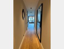 Langer, schmaler Flur in einer modernen Wohnung mit hellem Holzboden, beigen Wänden und Decken-Spotlights. Rechts hängt ein großer Spiegel, links eine runde Wandspiegel-Dekoration; am Ende führen Glastüren zu einem Wohnbereich mit Blick nach draußen.