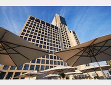 Ein modernes Hochhaus mit einer hellen Fassade und zahlreichen Fenstern, aufgenommen von unten. Im Vordergrund befinden sich große Sonnenschirme und eine Außenterrasse mit einer Bar.