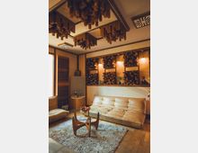 Wohnzimmer mit modernem Design, ausgestattet mit beigen Ledersofas, einem runden Glastisch auf einem flauschigen Teppich und stilisierten Holzelementen an Decke und Wänden. Die Beleuchtung erfolgt durch warme Wandlampen, und einzelne Dekorationsobjekte befinden sich auf Regalen.