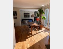 Esszimmer in einer modernen Wohnung mit Holzboden, einem rechteckigen Holztisch und mehreren Stühlen. Große Fenster rechts lassen Tageslicht hinein, und im Hintergrund ist ein Wohnzimmer mit Sofa und Wandkunst sichtbar.