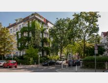 Mehrstöckiges Wohnhaus in einer grünen Straßenecke, teils mit Efeu bewachsen, umgeben von Bäumen und parkenden Autos. Im Vordergrund Straßenlaternen, Verkehrsschilder und Gehwege.