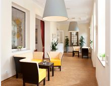 Heller Aufenthaltsraum mit großen Fenstern, gelben Polstermöbeln, Holzboden und Pflanzen. Im Hintergrund hängen moderne Lampen, und es gibt dekorative Glasfenster sowie eine Tür mit roten Vorhängen.