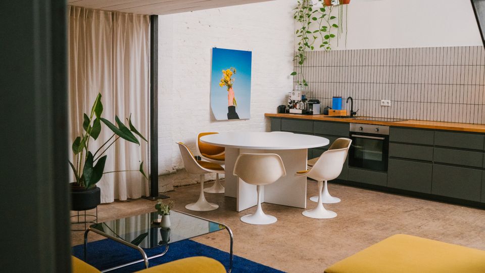 Offene Wohnküche mit weißer Backsteinwand, langer Küchenzeile mit dunklen Schränken und Holzarbeitsplatte sowie hell gefliestem Spritzschutz. In der Mitte steht ein ovaler weißer Esstisch mit vier Stühlen; daneben hängen Pflanzen von der Decke, ein Vorhang trennt den Raum und im Vordergrund liegt ein blauer Teppich mit Glastisch.