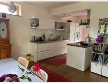 Moderne Küche und Essbereich in einem offenen Raum mit weißem Mobiliar, Holzfußboden und roten Akzenten. Links steht ein Holzschrank, daneben ein Esstisch mit Blumen, rechts eine Küchenzeile mit Spüle, Herd und offenen Regalen.