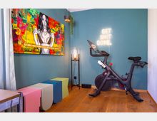 Ein kleiner Fitnessraum mit einer türkisfarbenen Wand, an der ein buntes, modernes Kunstwerk hängt und ein leuchtender Neon-Schriftzug „GET SHIT DONE“ angebracht ist. Im Raum stehen ein schwarzes Spinning-Bike, bunte Würfelhocker und eine Lampe mit Glaskuppel auf einem schwarzen Ständer.
