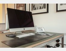 Moderner Arbeitsbereich mit einem großen Computerbildschirm, einer Tastatur, einem Taschenrechner und einem Festnetztelefon auf einem Schreibtisch. An der weißen Wand im Hintergrund hängen gerahmte Schwarz-Weiß-Fotografien.