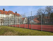 Eingezäunter Sportplatz mit rotem Kunststoffbelag und Basketballkorb, umgeben von Grasfläche und kahlen Bäumen. Im Hintergrund stehen mehrstöckige Gebäude mit roten Ziegeldächern sowie ein Hochhaus.