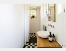 Schmales Badezimmer mit weiß gestrichenen Wänden, schwarzem und weißem Schachbrettboden, einem Waschbecken auf einer Holzplatte, einer Wandarmatur sowie einem kleinen Fenster mit Gitter und Pflanzen darauf.