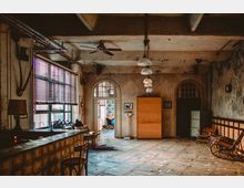 Ein Raum in einem industriellen Vintage-Stil mit freiliegenden Deckenbalken, gemustertem Fliesenboden und großen Fenstern mit Jalousien. Möbel wie eine Holzbar, Stühle, ein Schrank und ein Schaukelstuhl ergänzen die Einrichtung; im Hintergrund ist eine Tür mit Rundbogen und gemustertem Textil sichtbar.