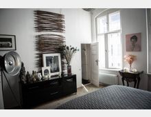 Schlafzimmer mit heller Wandfarbe und Dekorationen. Links steht eine schwarze Kommode mit Skulpturen, Kerzen und einem gerahmten Bild, daneben eine große Fotolampe; darüber hängen künstlerische Holzdekorationen. Rechts befindet sich ein Fenster mit einem Heizkörper darunter und ein kleiner Beistelltisch mit Trockenblumen und Kerzen.