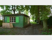 Grüne Holzlaube mit Fenstern und grünen Fensterläden, umgeben von hohen Bäumen und einer Wiese im Hintergrund. Der Boden ist größtenteils erdig, mit vereinzeltem Grasbewuchs, und ein kleiner Weg führt entlang der Seite des Gebäudes.