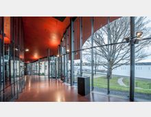 Modernes Foyer mit geschwungenem roten Dach und bodentiefen Glasfenstern, die den Blick auf eine Uferlandschaft mit Bäumen, Gehwegen und Wasser freigeben. Die Innenraumgestaltung wirkt offen, mit Lichtinstallationen und glatten roten Böden.