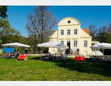 Gelbes, zweistöckiges Gebäude im klassizistischen Stil mit Rundgiebel, davor eine Wiese mit zahlreichen Tischen und Stühlen unter Sonnenschirmen. Im Hintergrund sind Bäume und ein blauer Himmel sichtbar, die Umgebung wirkt parkähnlich.