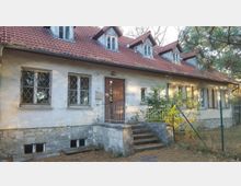 Ein eingeschossiges Gebäude mit verputzter Fassade, teils vergitterten Fenstern, einem roten Ziegeldach mit mehreren Dachgauben und einer kleinen Eingangstreppe mit Metallgeländer. Im Vordergrund befindet sich ein Gartenbereich mit Bäumen und einem Maschendrahtzaun.