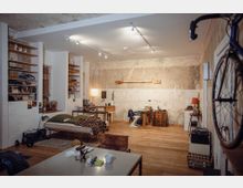 Wohnraum im industriellen Stil mit sichtbaren Betonwänden und Holzboden. Der Raum enthält ein einfaches Bett, Regale, einen Schreibtisch, verschiedene Möbelstücke und eine an der Wand montierte Fahrradhalterung.