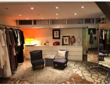 Ein eleganter Ankleideraum mit Kleiderstangen voller bunter Kleidung an beiden Seiten, einem weißen Sideboard mit Deko-Objekten und Bildern, sowie zwei lila Sesseln auf einem Teppich. Der Boden besteht aus ungleichmäßigen Steinplatten, und ein großer Spiegel steht an der rechten Seite.