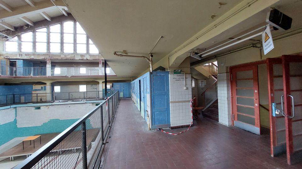 Ein verlassenes Hallenbad mit leerem Becken, blauer und weißer Fliesenverkleidung sowie hohen, bogenförmigen Fenstern. Im Vordergrund befindet sich ein Flur mit blauen Umkleidekabinen, einer Treppe und abgesperrtem Bereich.
