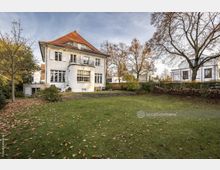 Zweistöckiges weißes Haus mit rotem Ziegeldach, großen Fenstern und einem kleinen Balkon, umgeben von einem Rasen mit Herbstlaub und mehreren Bäumen. Im Hintergrund sind weitere Wohnhäuser und ein Gartenzaun erkennbar.