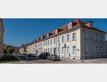 Straßenszene mit gepflegten, pastellfarbenen Wohnhäusern im barocken Stil, roten Ziegeldächern und symmetrischen Fassaden; geparkte Autos am Kopfsteinpflasterrand und ein klarer blauer Himmel im Hintergrund.